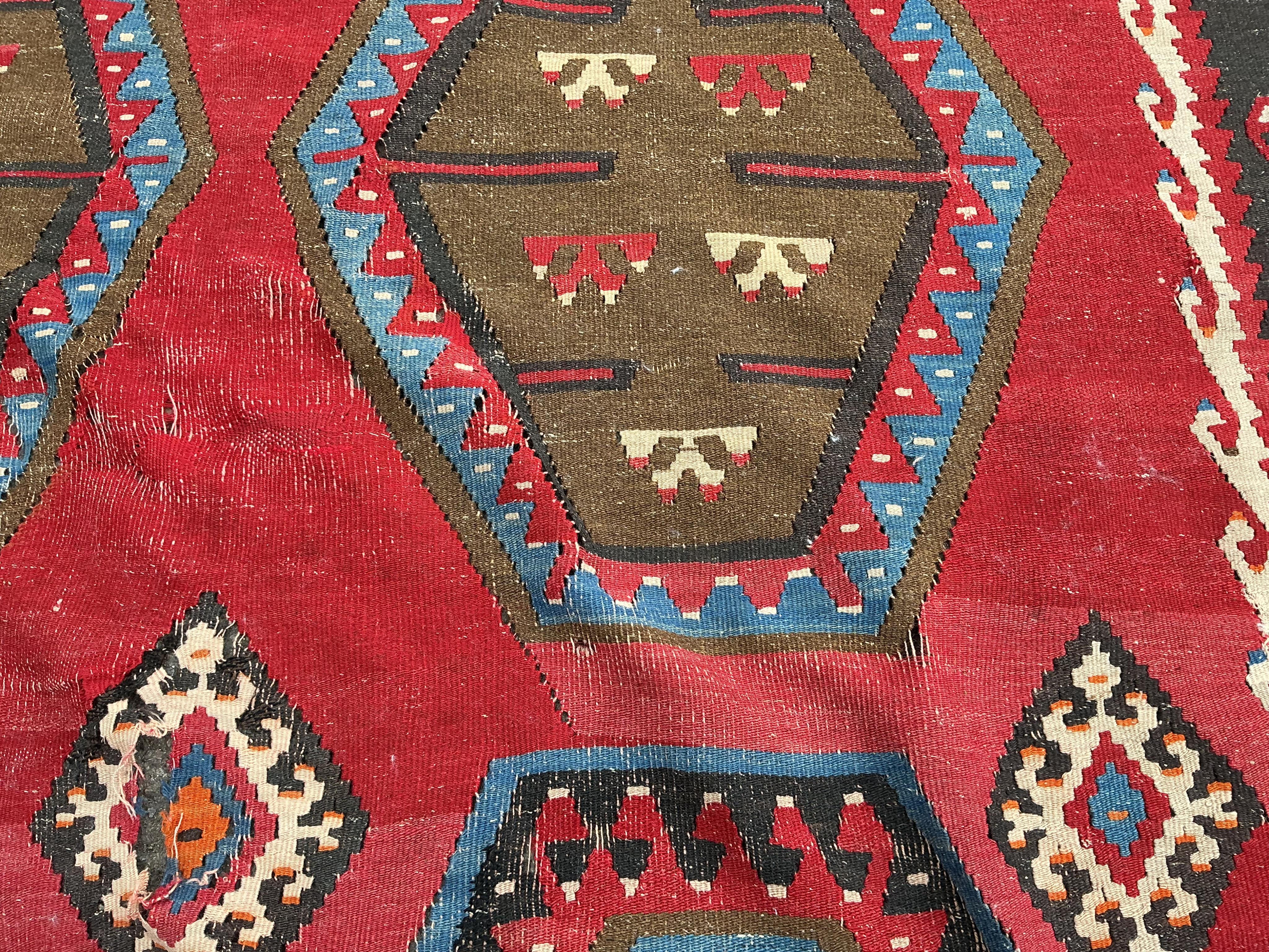 A Turkish Kilim flatweave polychrome carpet, woven with three rows of hexagons, on a red ground, (severely holed and damaged in places), 373 x 267cm
